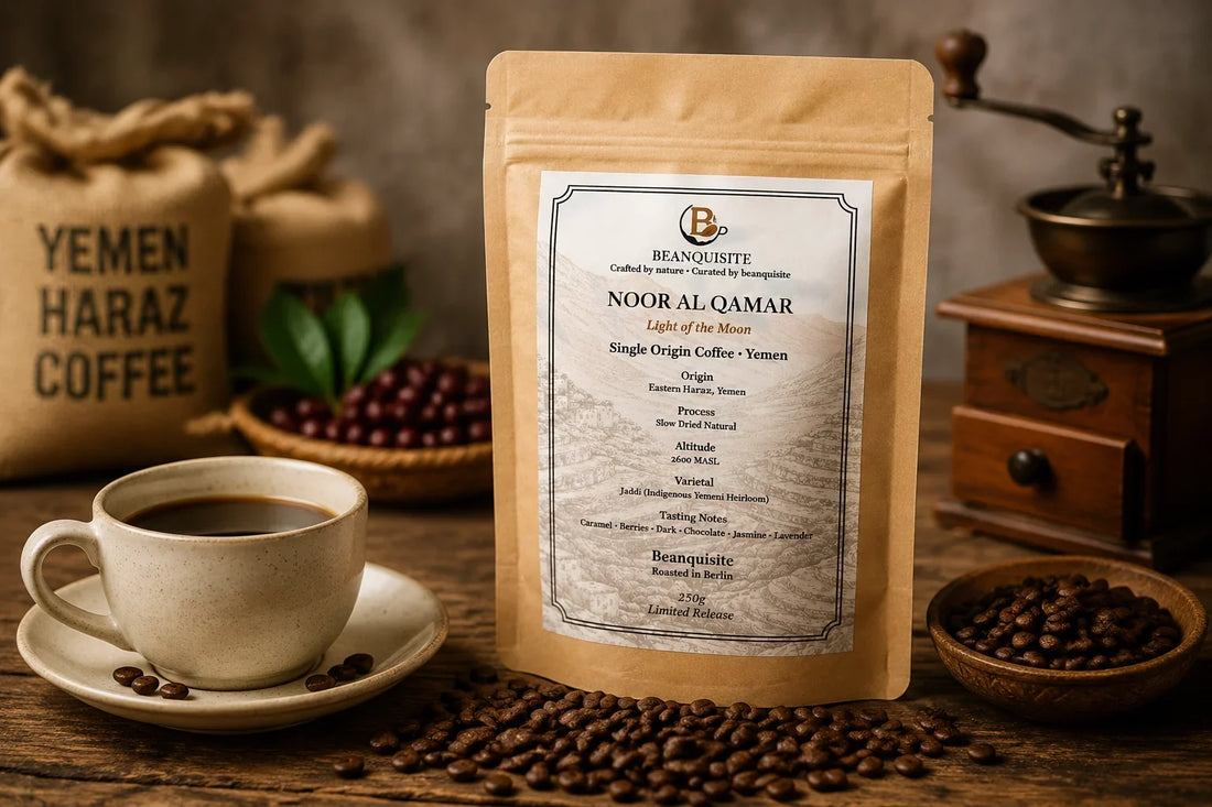 Beanquisite Noor Al Qamar Single Origin Yemeni coffee beans pouch displayed on a rustic wooden table with roasted coffee beans, a coffee cup, Yemen Haraz coffee sack, and traditional grinder in a warm natural setting.